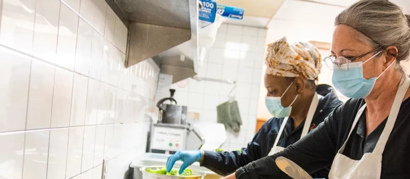 Deux personnes faisant des sandwichs dans une cuisine commerciale portant des masques et gants.