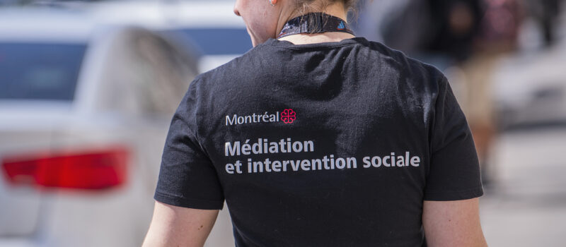 Photo d’une intervenante de l’équipe ÉMMIS prise de dos. Elle porte un t-shirt sur lequel on peut voir le logo de la Ville de Montréal et l’inscription « Médiation et intervention sociale ».