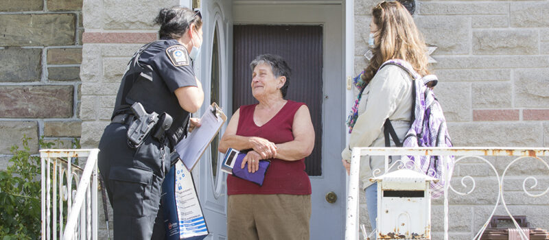 Une policière du Service de police de la Ville de Montréal et une intervenante discutent avec une dame souriante sur le pas de la porte d’une maison. Crédit photo : Véronique Lussier, SPVM.