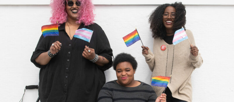 Trois femmes agitent chacune le drapeau arc-en-ciel LGBTQ2+ et le drapeau transgenre en souriant . Deux sont debout et l’une est assise dans un fauteuil roulant.