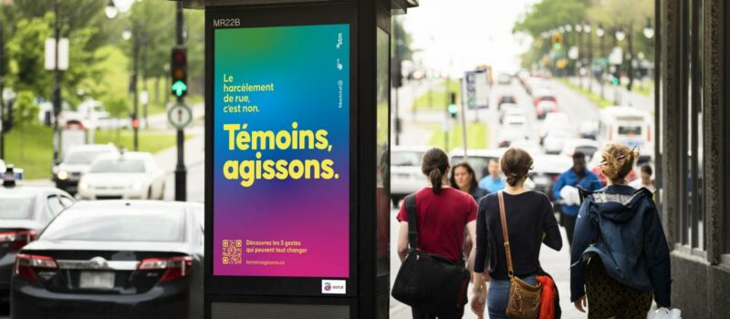 Une colonne affiche : “Le harcèlement de rue, c’est non. Témoins agissons.” Trois personnes marchent de dos à droite de la colonne, dans un contexte urbain animé.