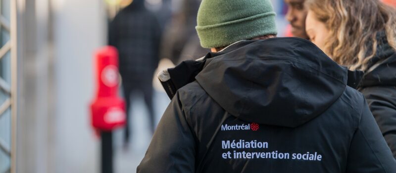 Personne, vue de dos, portant une tuque verte et un manteau noir avec l’identification visuelle de la Ville de Montréal et la mention « médiation et intervention sociale ». Cette personne est dans une rue où d’autres personnes se trouvent en arrière-plan.