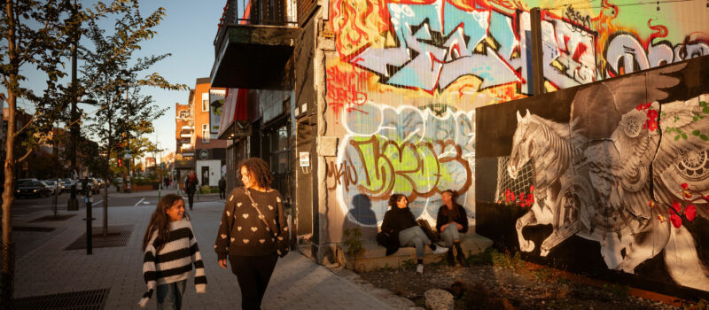 Femme et fille qui marchent souriantes sur une rue montréalaise au coucher de soleil. Deux femmes assises qui parlent en arrière-plan.