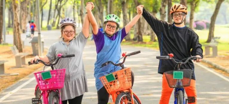 Trois enfants debout auprès de leur vélo dans un parc. Ils regardent vers l’objectif de la photo et se tiennent les mains dans un mouvement de célébration.