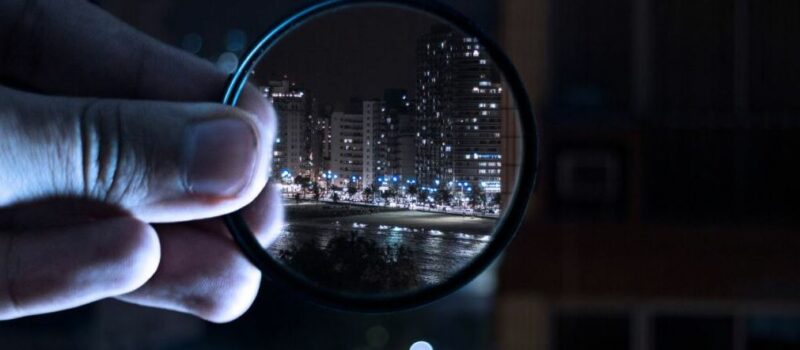 Une main tient une loupe devant un paysage urbain nocturne. À travers la loupe, la ville apparaît nette et lumineuse, tandis que le reste de l’image est flou et sombre.