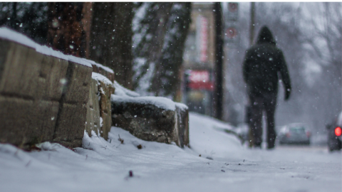 L'image montre une scène hivernale avec de la neige tombant doucement. Au premier plan, il y a un mur de pierre recouvert de neige. En arrière-plan, on peut voir une personne marchant sur un trottoir enneigé et flou, ainsi que des voitures en mouvement.