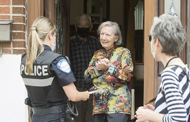 Un couple, composé d’un homme et d’une femme, se tient à la porte de leur domicile pour accueillir la visite d’une policière accompagnée d’une intervenante communautaire.