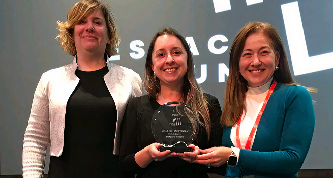 Trois femmes se tiennent debout sur une scène, la personne au centre tenant un trophée en verre transparent avec des inscriptions gravées sur sa surface. Elles sont habillées de manière formelle. La première personne à gauche porte une veste blanche sur une robe noire. La deuxième est vêtue tout en noir. La troisième porte un chandail bleu par-dessus un chandail blanc, avec un pantalon noir.