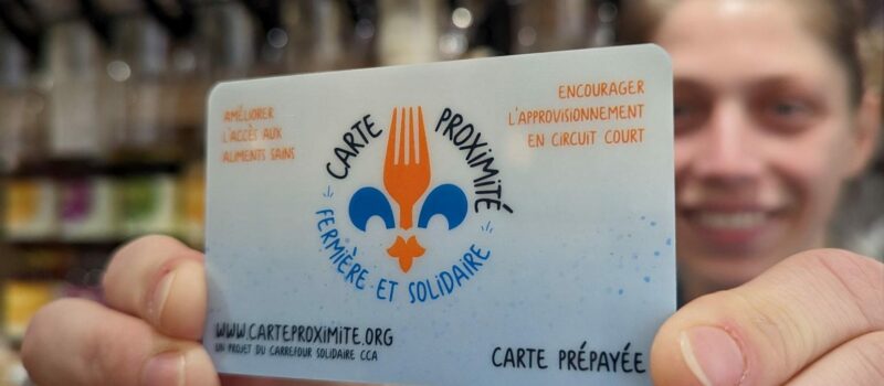 Une personne souriante tient en avant-plan une carte prépayée intitulée « Carte Proximité – Fermière et solidaire ». La carte affiche les slogans « Améliorer l’accès aux aliments sains » et « Encourager l’approvisionnement en circuit court ». En arrière-plan, on distingue des contenants d’aliments en vrac flous, suggérant un commerce local ou une épicerie solidaire.