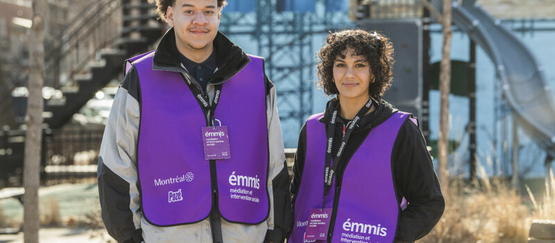 Un intervenant et une intervenante de l’ÉMMIS se tiennent debout côte à côte dans un parc. Derrière eux, une murale avec des échafaudages de couleur bleue. Les deux personnes portent une veste mauve avec les logos de la Coalition Pozé et de la Ville de Montréal. Il et elle sourient à la caméra et ont les deux mains dans les poches.
