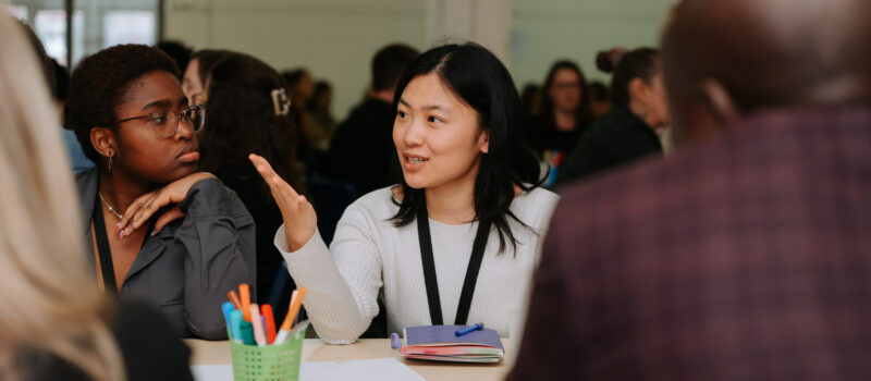 Un groupe de personnes est assis autour d’une grande table dans une grande salle. Deux personnes sont à l’avant-plan. Une personne, placée au centre, porte un pull blanc et semble parler ou expliquer quelque chose, en utilisant ses mains pour accompagner ses paroles. Les autres personnes autour écoutent attentivement ou échangent entre elles. À l’arrière-plan, on voit encore plus de personnes assises et en discussion, ce qui donne l’impression d’un environnement collaboratif, comme un atelier ou une séance de travail en groupe.