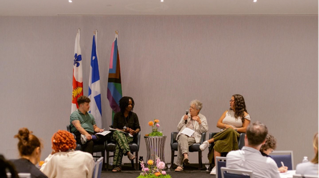 Un panel constitué de quatre personnes assises côte à côte sur une scène. De gauche à droite : Henri-June Pilote, Sheba Akpokli, Line Chamberland et Géorgie Gagné. Derrière elles, on voit trois drapeaux : celui de la Ville de Montréal, celui du Québec et celui de la Fierté. Des arrangements floraux décorent le devant de la scène. Dans la salle, des personnes sont assises et écoutent attentivement le panel.