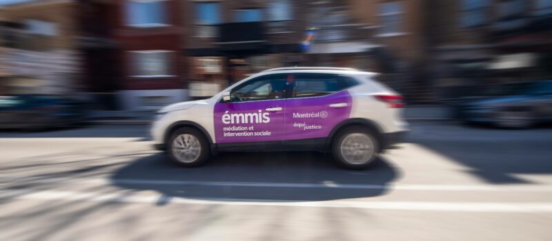 Au centre de l'image, une voiture de l'ÉMMIS est en mouvement sur une rue de Montréal. La voiture est un VUS blanc et sur ses portières mauves, on voit le logo ÉMMIS - Médiation et intervention sociale, en plus de ceux de la Ville de Montréal et de l'organisme Équijustice. À l'arrière-plan, on aperçoit des appartements flous.