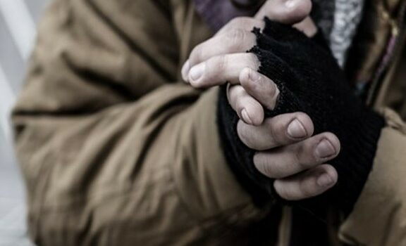 Gros plan sur des mains serrées dans des gants usés. La personne porte un manteau épais aux tons bruns et un vêtement de laine, suggérant un temps froid.