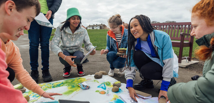 Groupe de jeunes participant à une activité extérieure, rassemblés autour d’une grande carte colorée posée au sol. Un ciel nuageux est visible à l’arrière-plan.